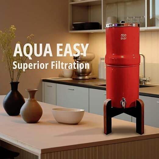 A red AquaEasy Stainless Steel Gravity Water Filter System, featuring a wooden stand, stainless steel tap and lid, displayed on a kitchen counter with vases and a bowl