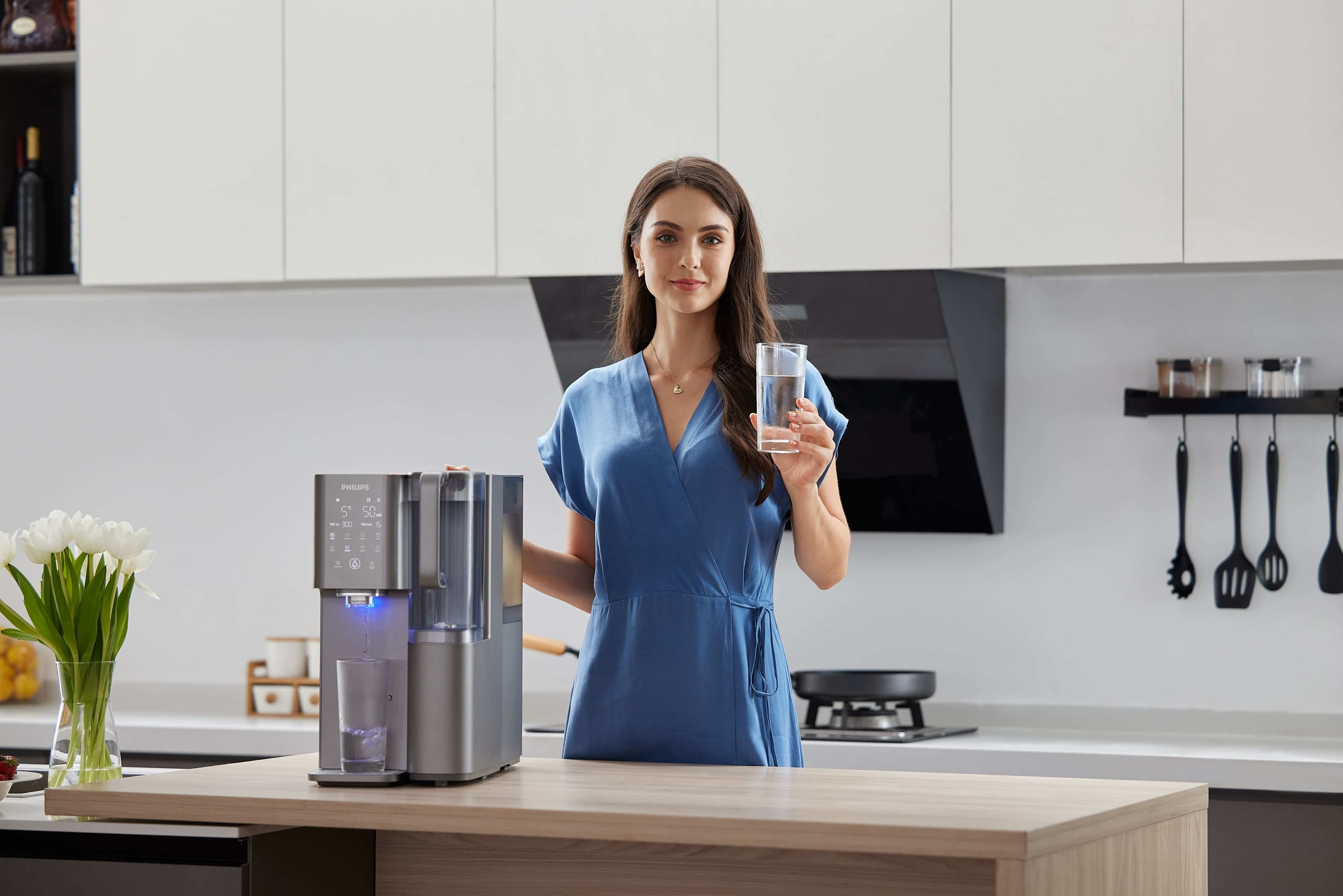 A woman in a blue dress sips water near a sleek Philips Aquaporin Reverse Osmosis Water Station with compressor cooling (model ADD6921DG/79) in a modern kitchen.
