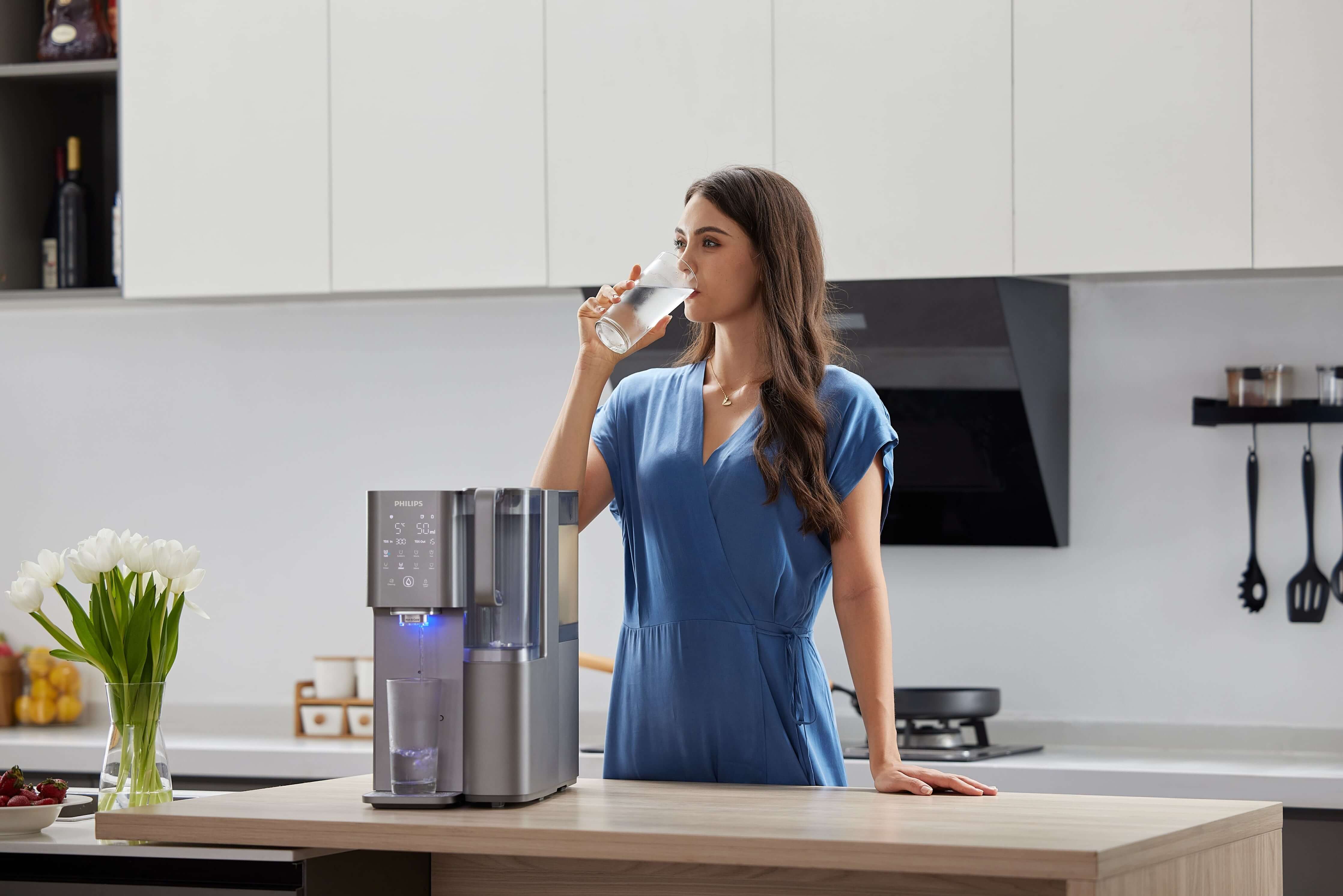In her modern kitchen, a woman in a blue dress savors a refreshing glass of water from the sleek Philips Aquaporin Reverse Osmosis Water Station (model ADD6921DG/79), featuring advanced compressor cooling and UV-C LED freshness technology.