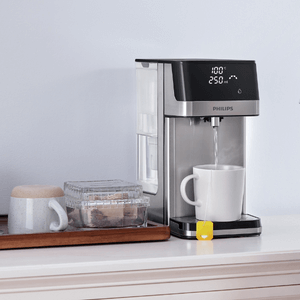 A Philips Compact Water Station ADD5910M/79 with Micro X-Clean Filtration dispenses instant hot water into a mug placed next to a tray holding another mug and a container of cookies.