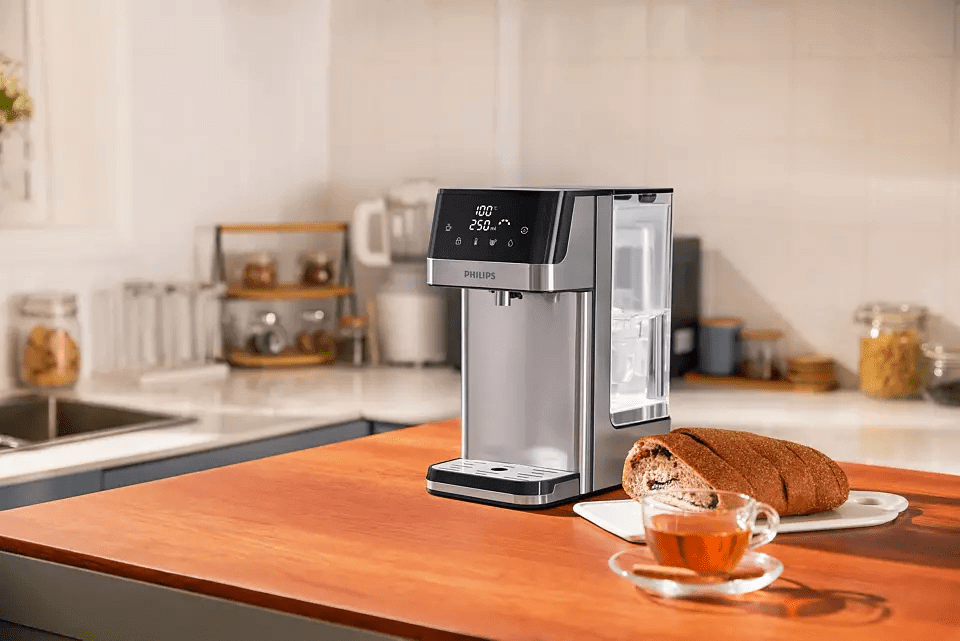 In a sleek kitchen setting, a Philips Compact Water Station ADD5910M/79 with Micro X-Clean Filtration rests on the counter beside a sliced loaf and cup of tea, providing instant hot water for convenience.