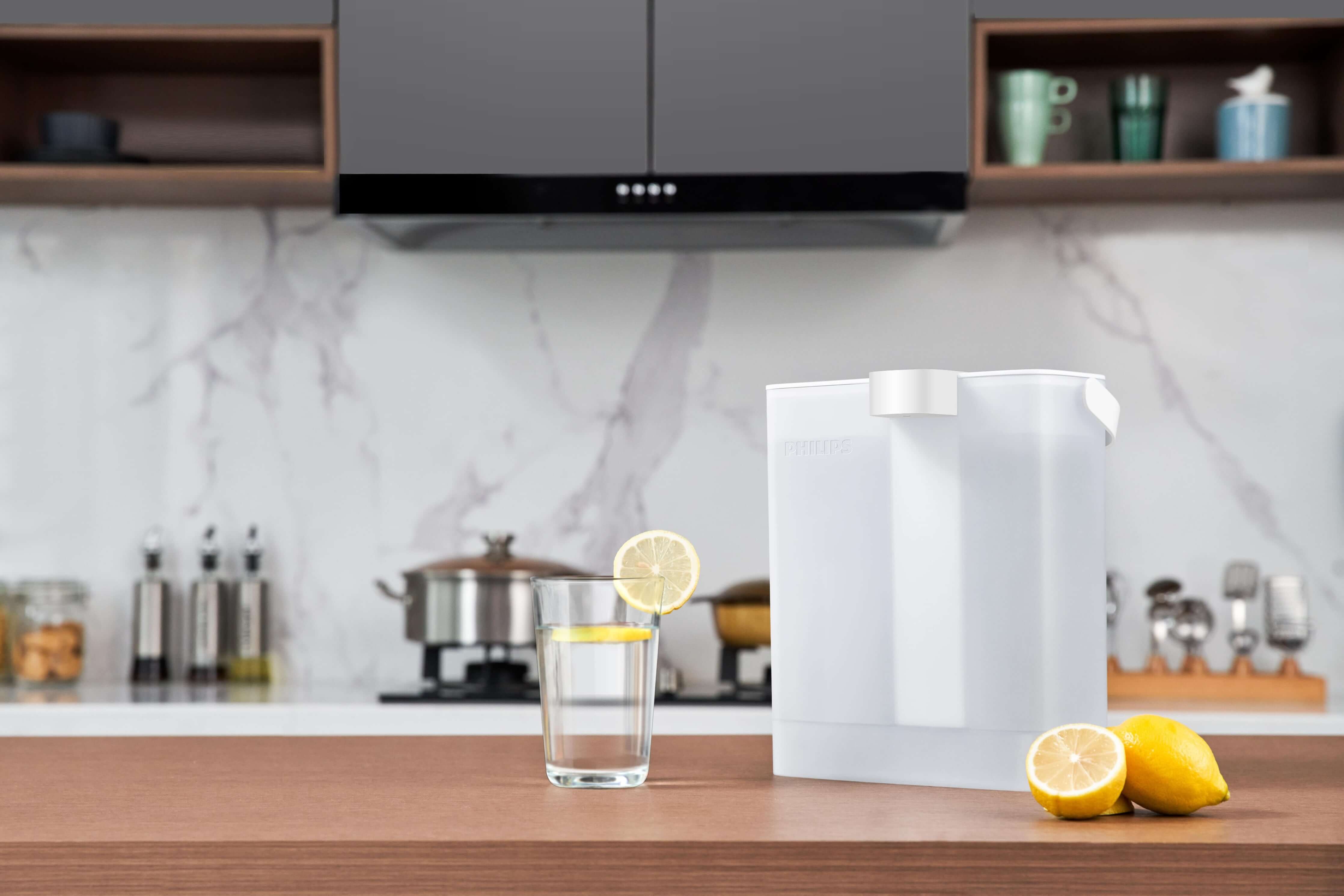 A Philips Next-generation instant water filter AWP2980WH with Micro X-Clean instant filtration technology and a glass with lemon slices are placed on the kitchen counter, with a stove and cookware visible in the background.