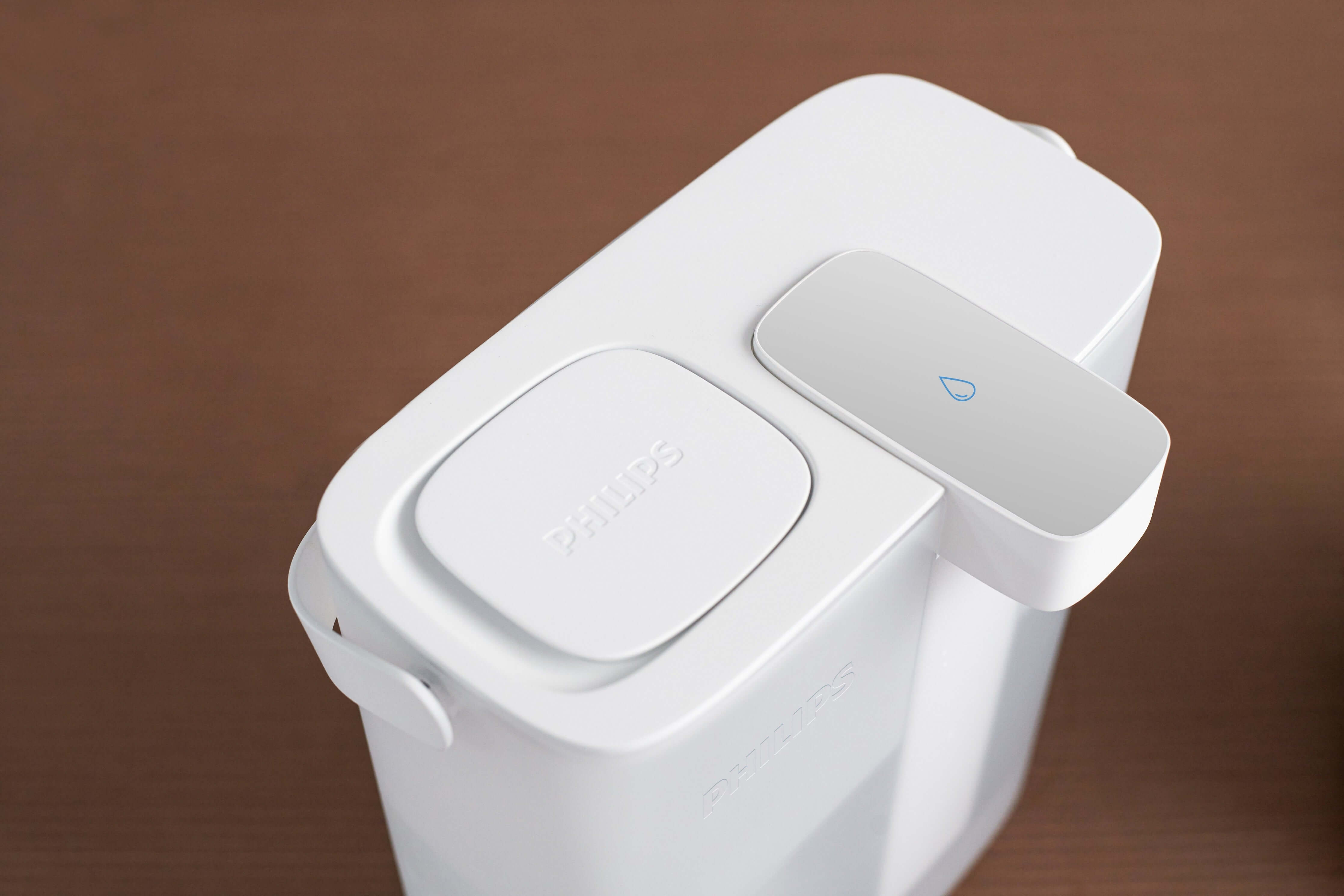The Philips Next-generation instant water filter AWP2980WH, featuring Micro X-Clean instant filtration technology and equipped with a top lid and spout, rests on a brown surface.