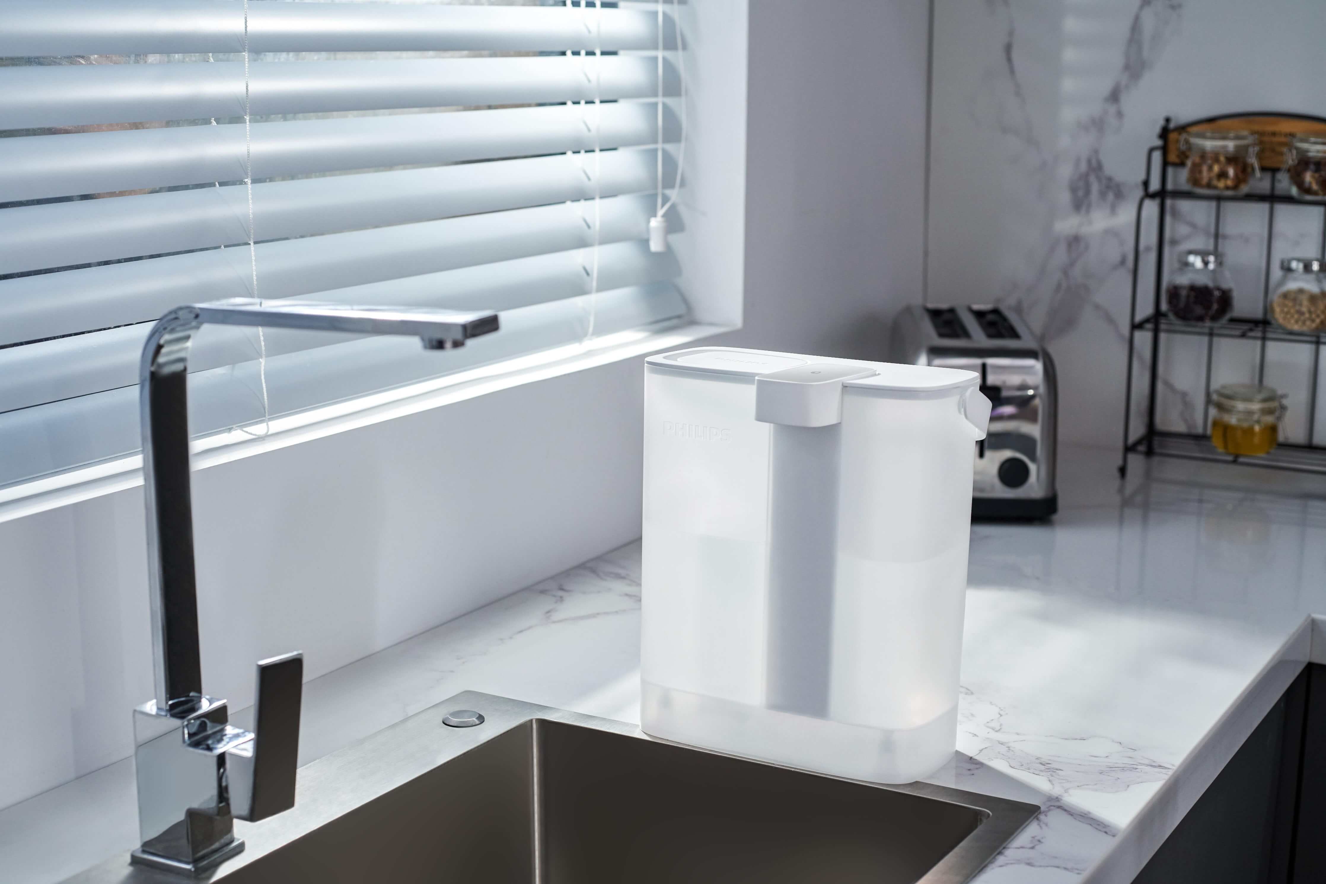 This contemporary kitchen boasts a Philips Next-generation instant water filter AWP2980WH with Micro X-Clean instant filtration technology on the counter, elegantly placed next to the sink and beneath a window with blinds.
