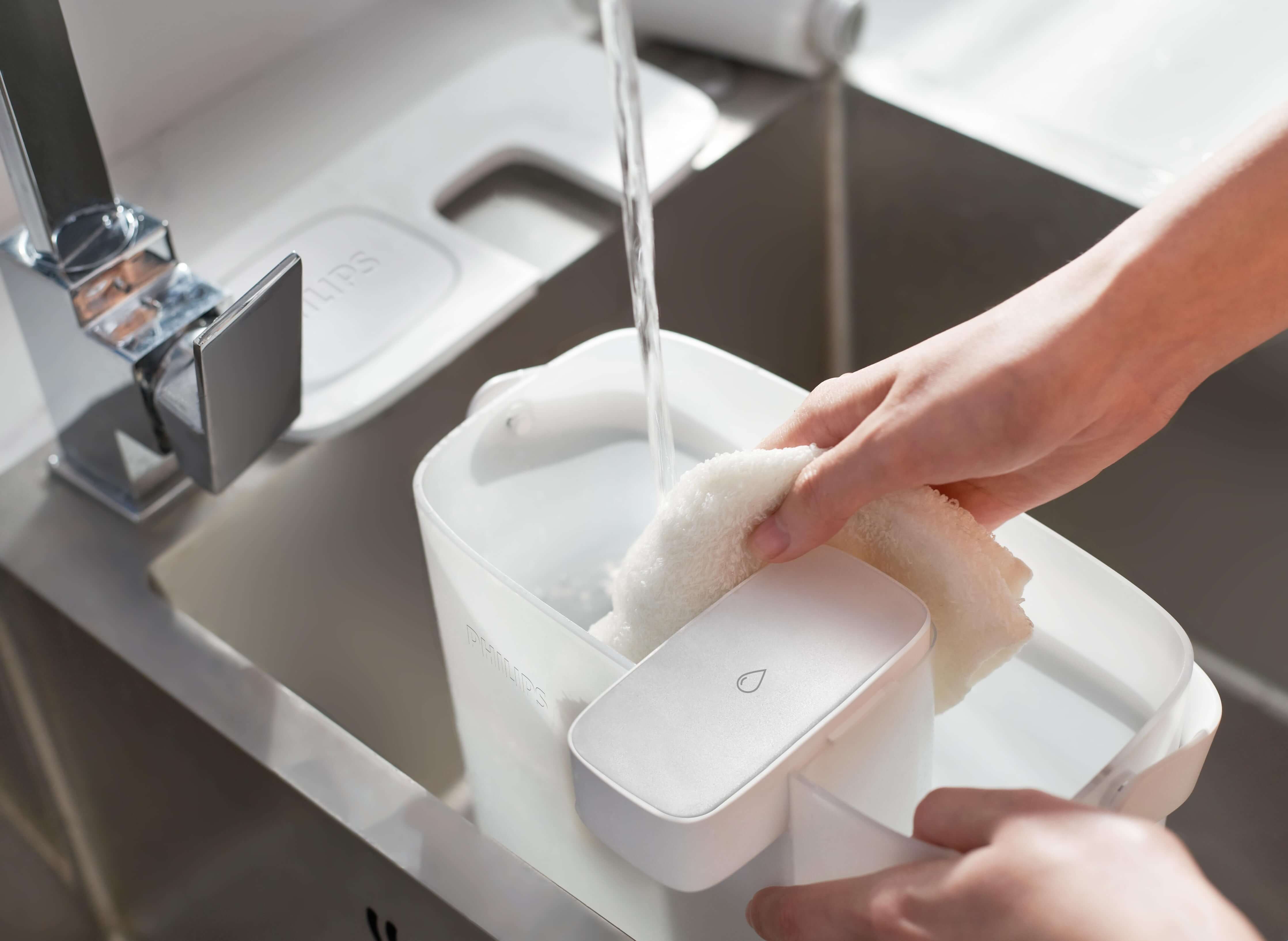 Hands washing a white container with a cloth under running water in a kitchen sink, utilizing the Philips Next-generation Instant Water Filter AWP2980WH with Micro X-Clean instant filtration technology for optimal cleanliness.