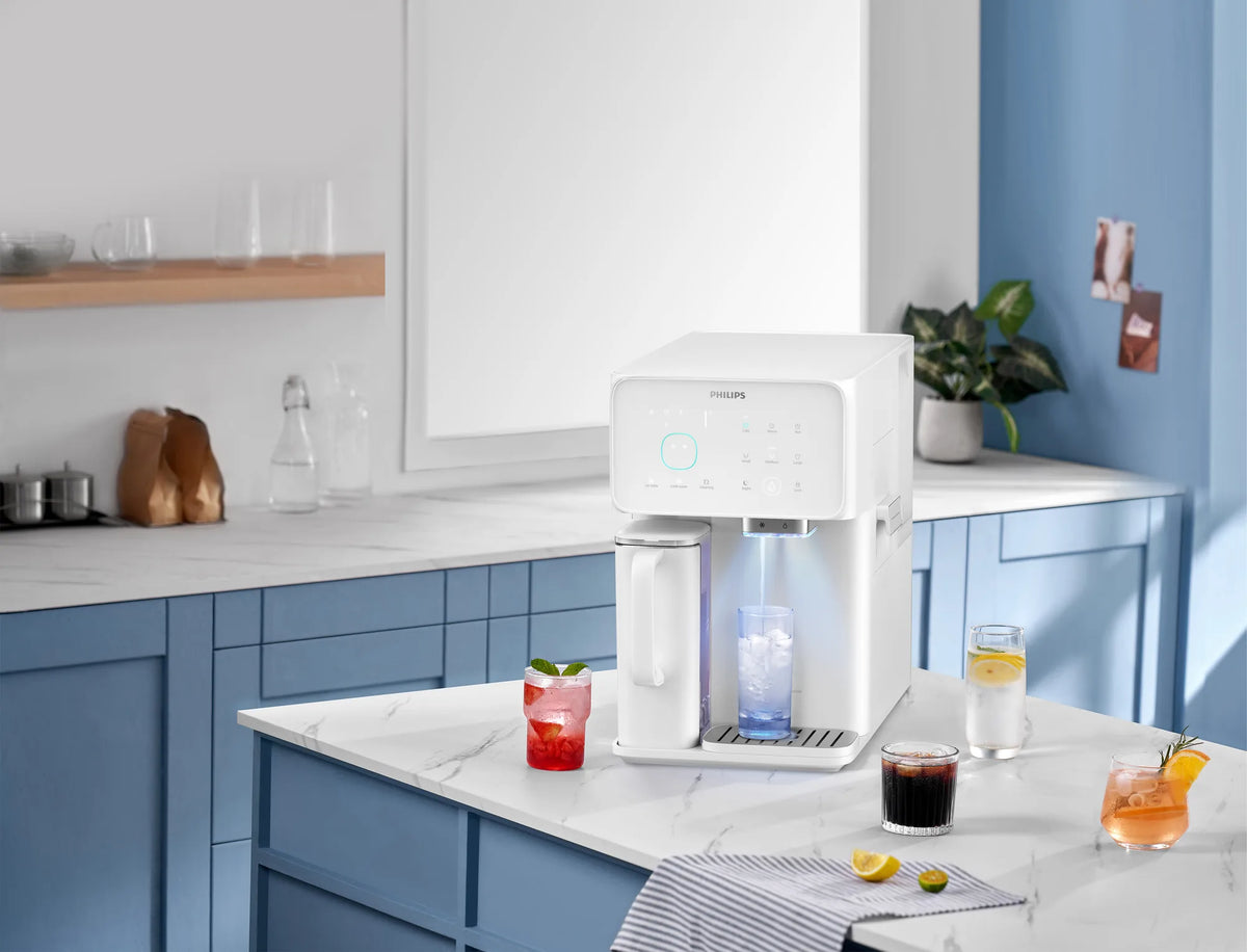 White Philips water station dispensing cold water into a glass of ice on a marble counter, surrounded by iced drinks and lemon slices in a blue kitchen.