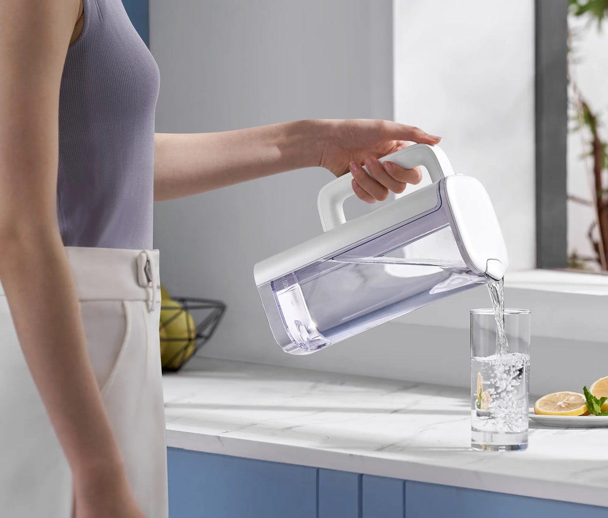 Woman in a lavender top pouring filtered water from the white Philips jug into a glass beside lemon slices on a marble counter.