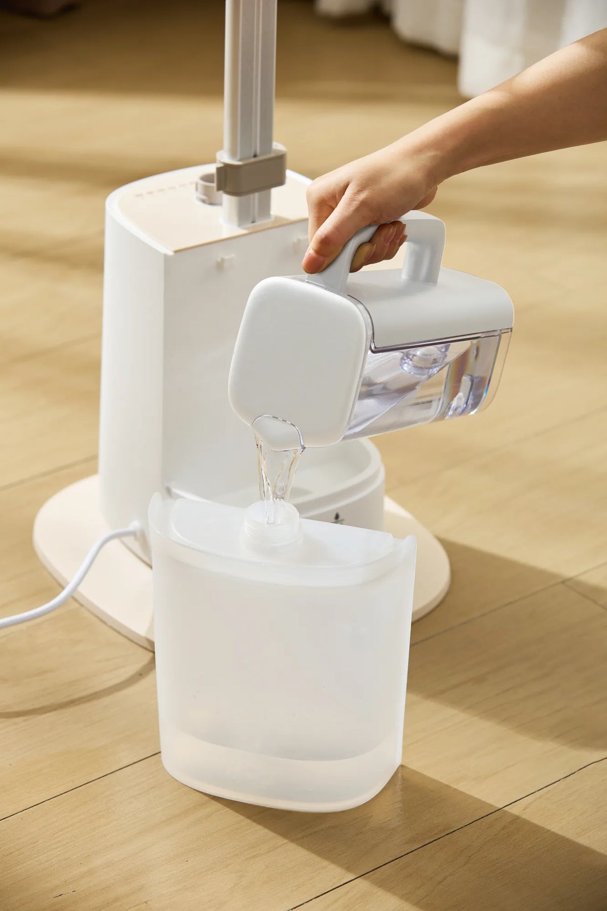 Hand pouring water from the white Philips water jug into the tank of a garment steamer on a wooden floor.