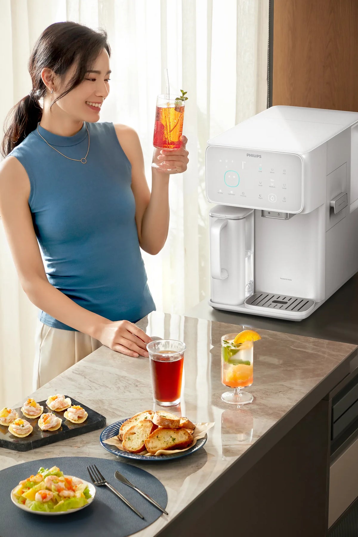 Smiling woman holding an iced drink beside the white Philips water station, with tea, bread, and appetizers on the counter.