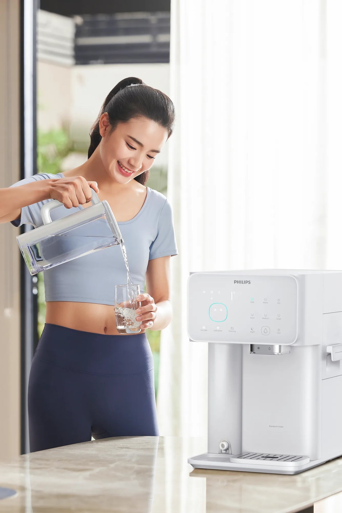 Smiling woman in athletic wear pouring filtered water from the Philips jug into a glass beside the white water station.