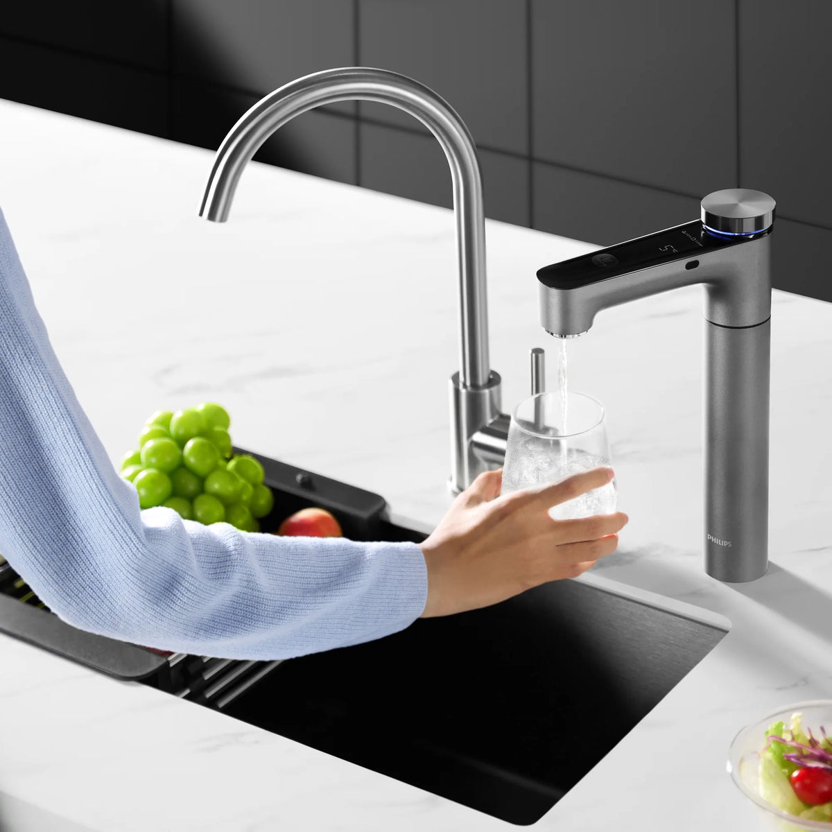 Hand holding a glass under the Philips RO tap as it dispenses chilled water, beside a stainless steel kitchen faucet and a sink with grapes.