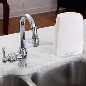 A chrome kitchen faucet is elegantly positioned on a marble countertop alongside the Aquasana Countertop Drinking Water Filter (AQ-4000) in white, which features Claryum® filtration technology.