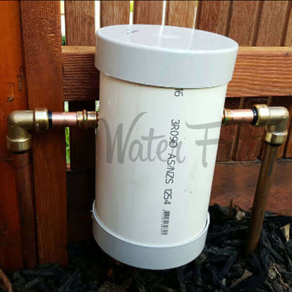 The cover to protect Grander Whole House System features a white cylindrical with brass connectors, mounted on a wooden fence.