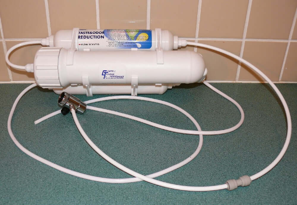 The 3 Stage Portable Reverse Osmosis Water Filter System, equipped with hoses, is placed on a green countertop against a tiled wall.