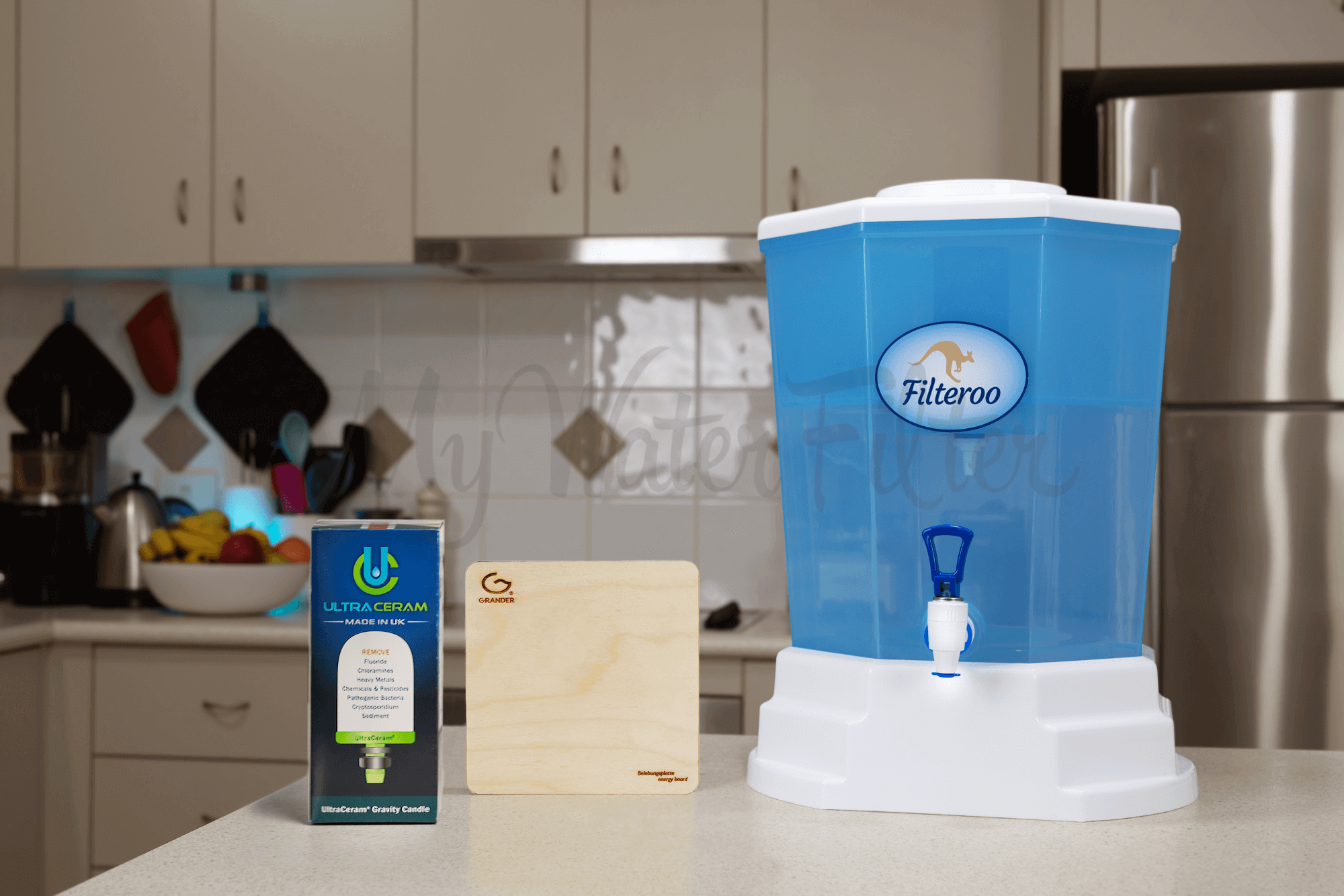 On the kitchen counter sits a Filteroo® Blue 20L Benchtop Gravity Water Filter with Ultraceram and Grander Board, accompanied by its box labeled "Ultraceram" and a wooden stand, set against a blurred background of kitchen items.