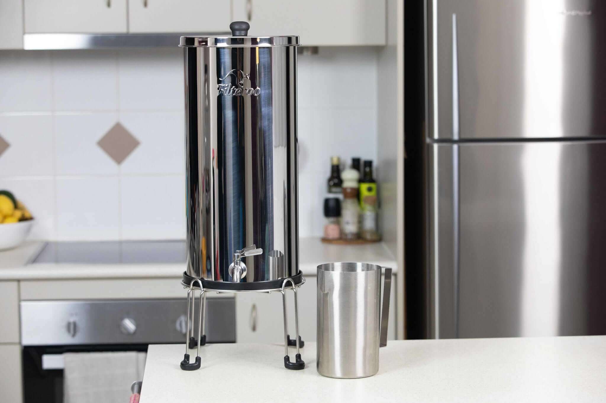 The Filteroo® Superoo 16L Stainless Steel Gravity Water Filter and stainless steel jug on a kitchen counter with appliances and decor in the background.