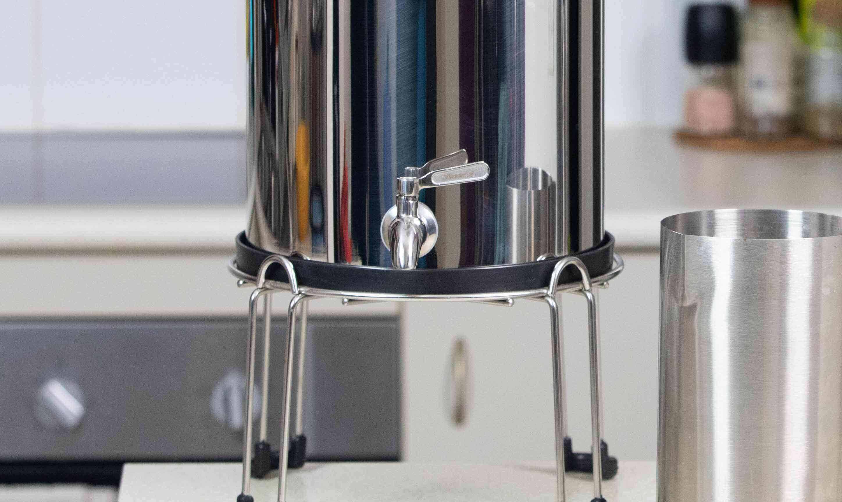 A Filteroo® Superoo 16L Stainless Steel Gravity Water Filter sits on a stand with a stainless steel tap, placed on a kitchen counter next to a jug.