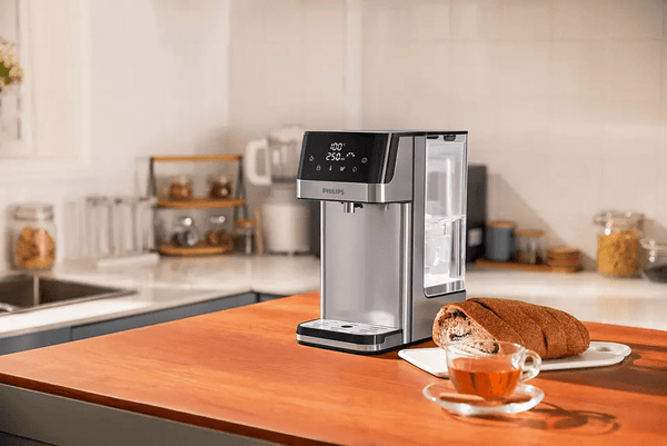 In a sleek kitchen setting, a Philips Compact Water Station ADD5910M/79 with Micro X-Clean Filtration rests on the counter beside a sliced loaf and cup of tea, providing instant hot water for convenience.