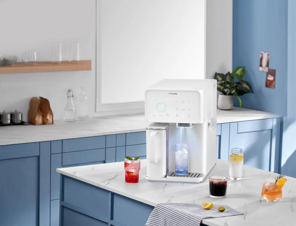White Philips water station dispensing cold water into a glass of ice on a marble counter, surrounded by iced drinks and lemon slices in a blue kitchen.
