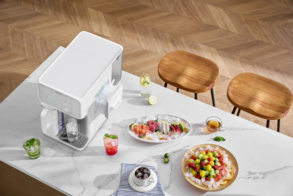 Overhead view of the white Philips water station with its ice drawer open, beside iced drinks, a sashimi platter, and a fruit bowl on a marble counter.
