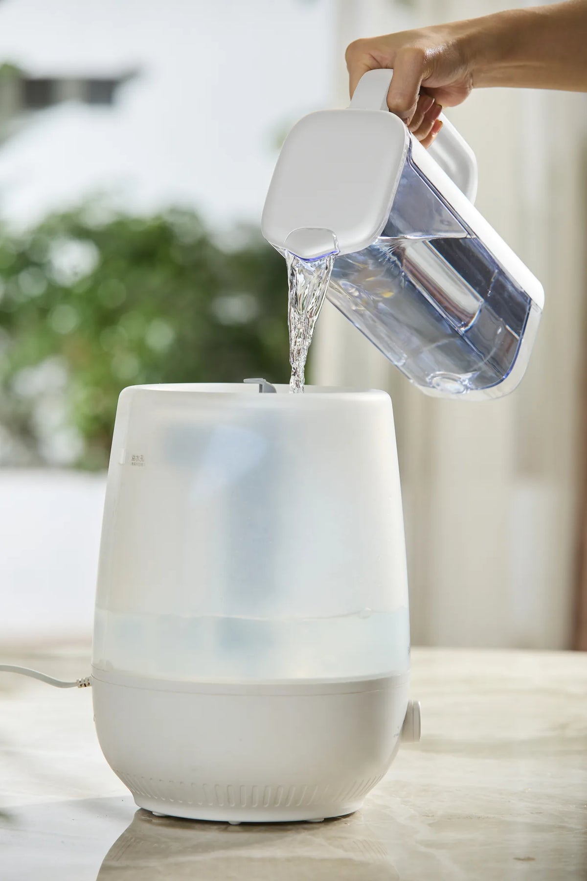 Hand pouring filtered water from the Philips jug into the tank of a white humidifier.