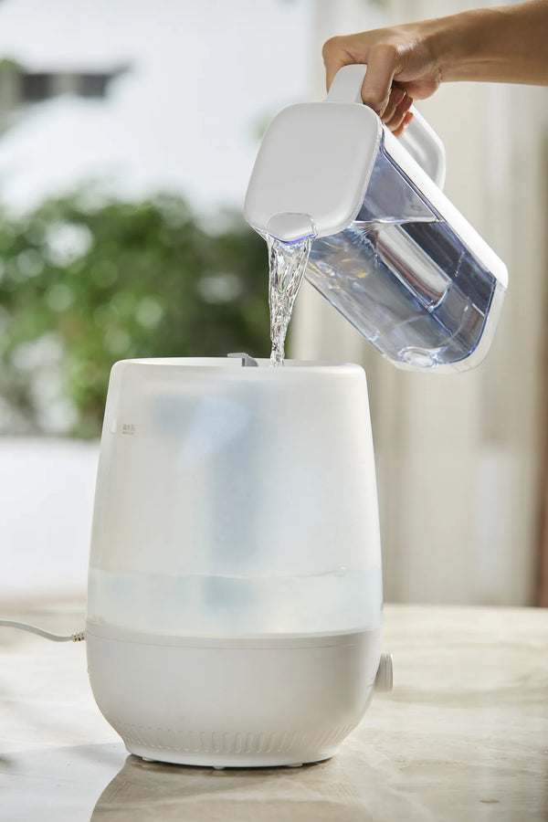 Hand pouring filtered water from the Philips jug into the tank of a white humidifier.