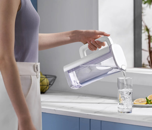 Woman in a lavender top pouring filtered water from the white Philips jug into a glass beside lemon slices on a marble counter.