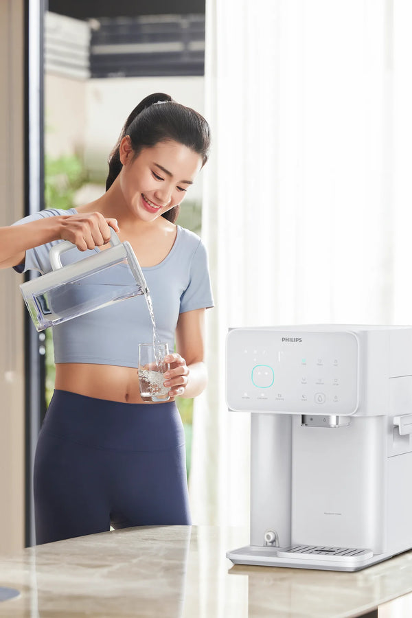 Smiling woman in athletic wear pouring filtered water from the Philips jug into a glass beside the white water station.