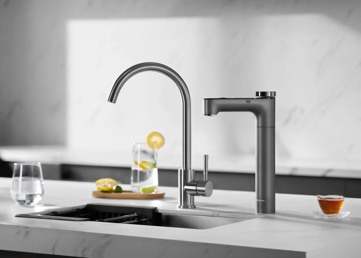 Philips RO tap beside a stainless steel kitchen faucet on a marble counter, with a glass of lemon water and a cup of tea nearby.