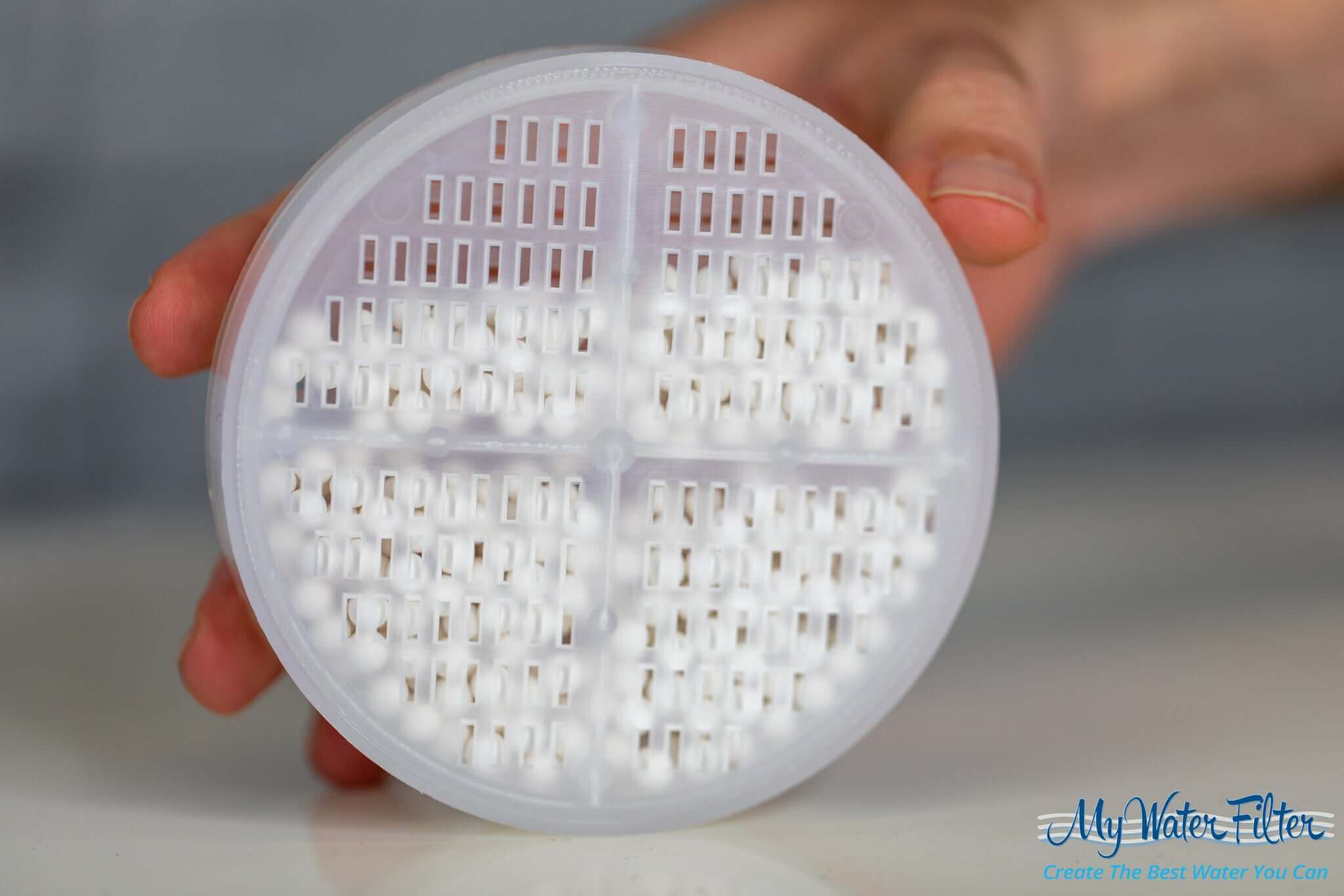 A hand holds a round white shower filter adorned with small holes, resting on a white surface. This product, known as the 