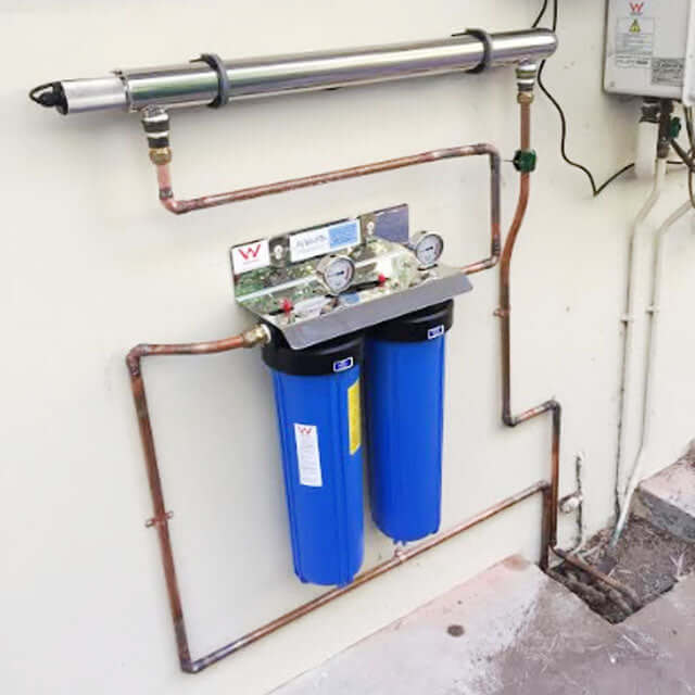 A My Water Filter system, the MWF 20" x 4.5" Twin Big Blue Whole House Rain Water Tank Filter System Complete with Ultraviolet Light, is mounted on the wall with dual blue filters and pressure gauges, seamlessly integrated into a whole house filtration setup.